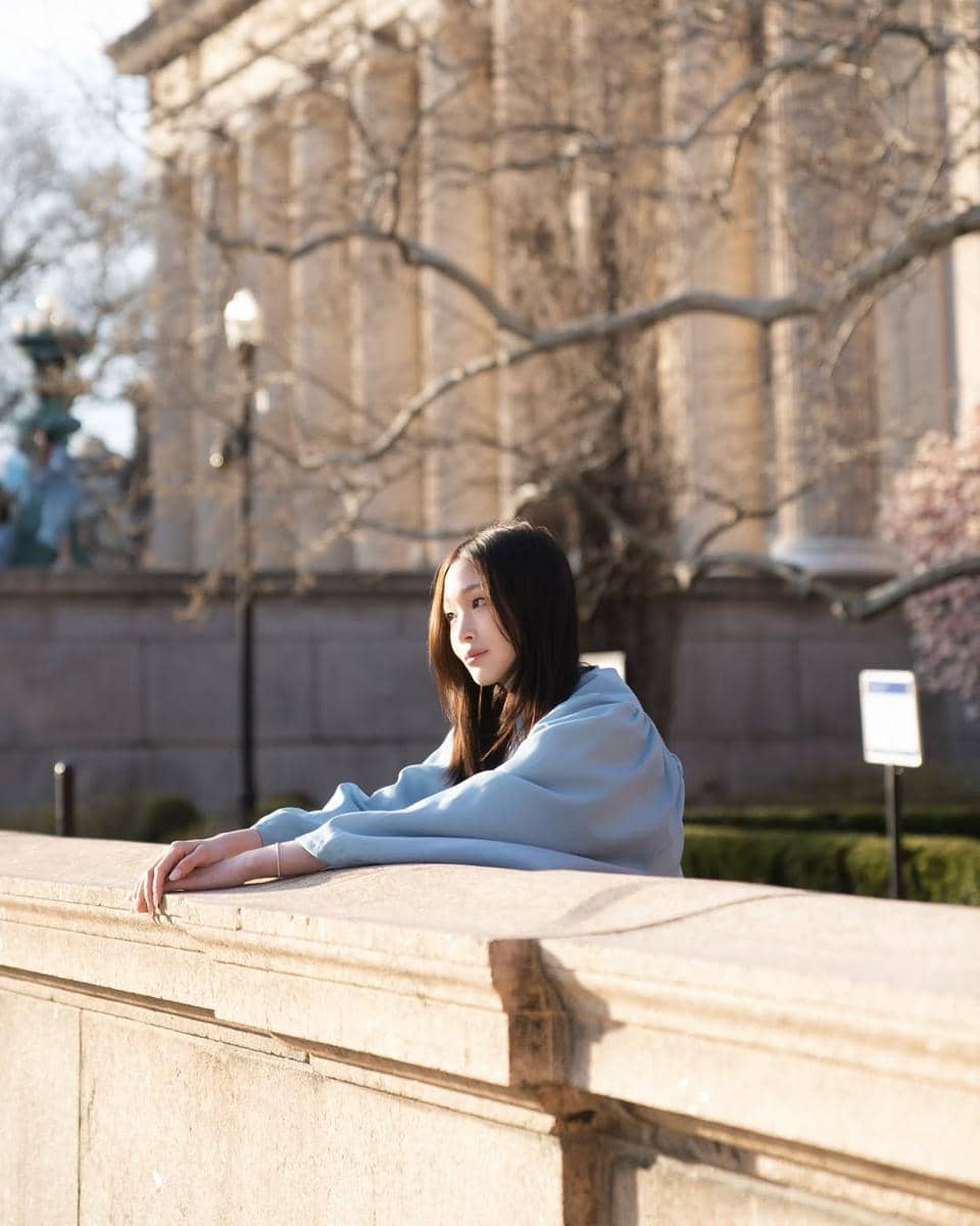 Seorang perempuan mengenakan jubah wisuda berwarna biru muda berdiri di dekat dinding batu dengan latar bangunan klasik di Columbia University.