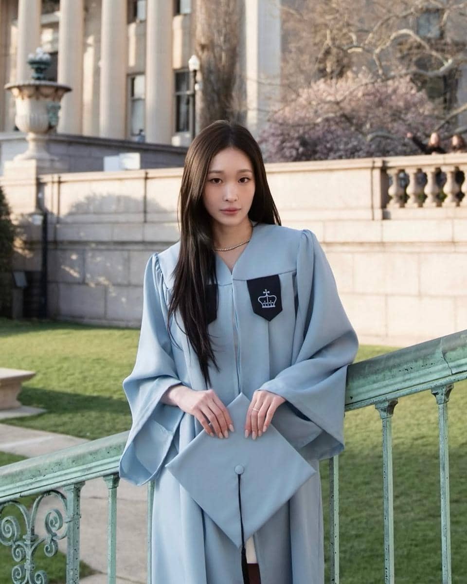 Seorang perempuan mengenakan toga wisuda berwarna biru muda berdiri di area kampus Columbia University dengan latar bangunan klasik.