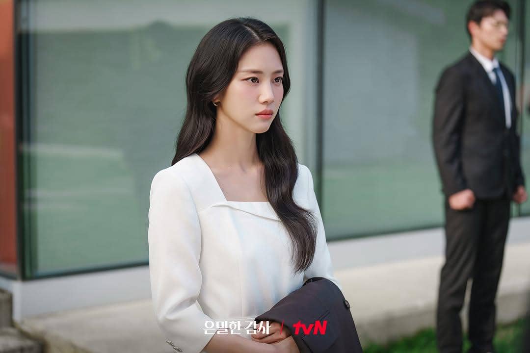 Seorang wanita berambut panjang mengenakan blus putih berdiri di luar gedung kaca dengan ekspresi serius, adegan dari drama Filing for Love.