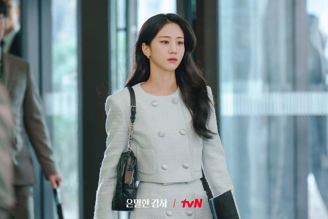 Seorang wanita mengenakan setelan jas abu-abu muda dengan tas hitam berjalan di area kantor dalam cuplikan drama Filing for Love.