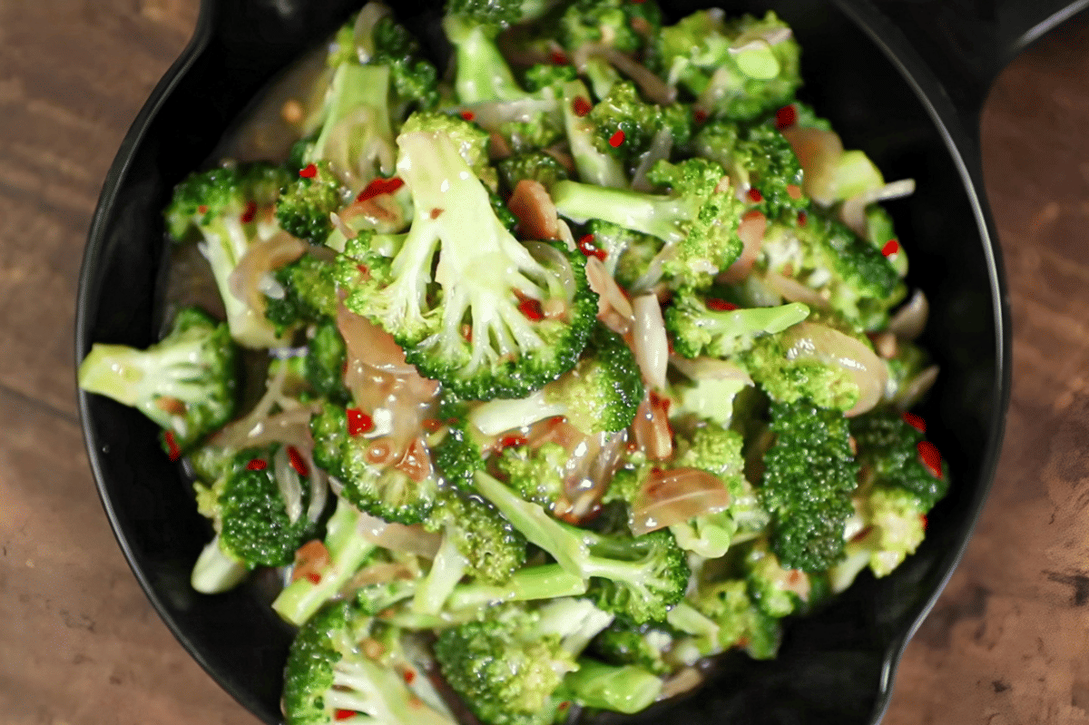 Salad brokoli Italia dengan potongan brokoli hijau segar, bawang, dan cabai merah disajikan dalam mangkuk hitam di atas meja kayu.