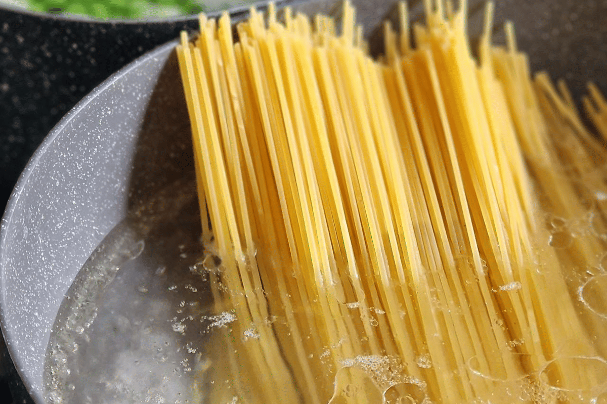 Pasta spaghetti kering sedang direbus dalam panci berisi air mendidih sebagai langkah awal membuat pasta e piselli.