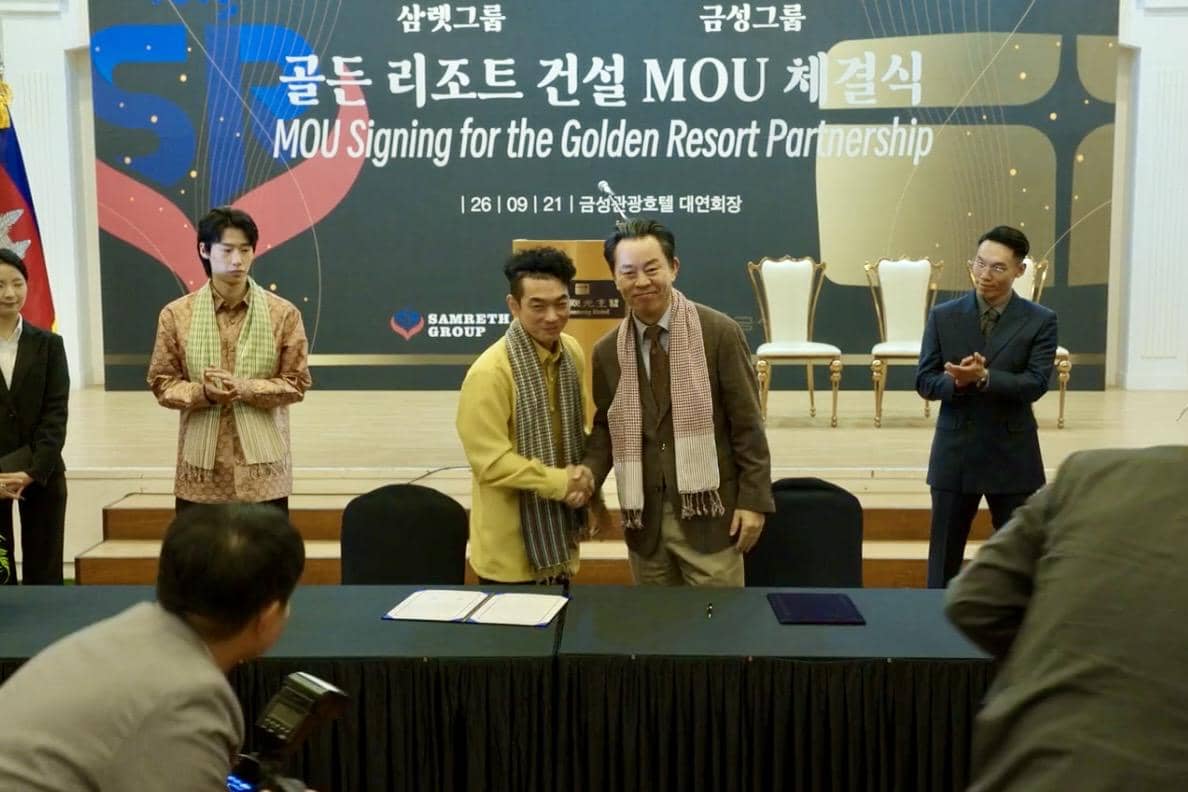 Beberapa orang berdiri di depan panggung saat acara penandatanganan MOU untuk kemitraan Golden Resort dalam cuplikan drakor Gold Land.
