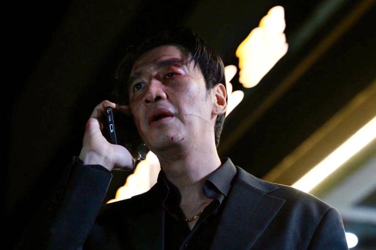Seorang pria berwajah lebam mengenakan setelan gelap sedang berbicara di telepon di bawah cahaya lampu malam.