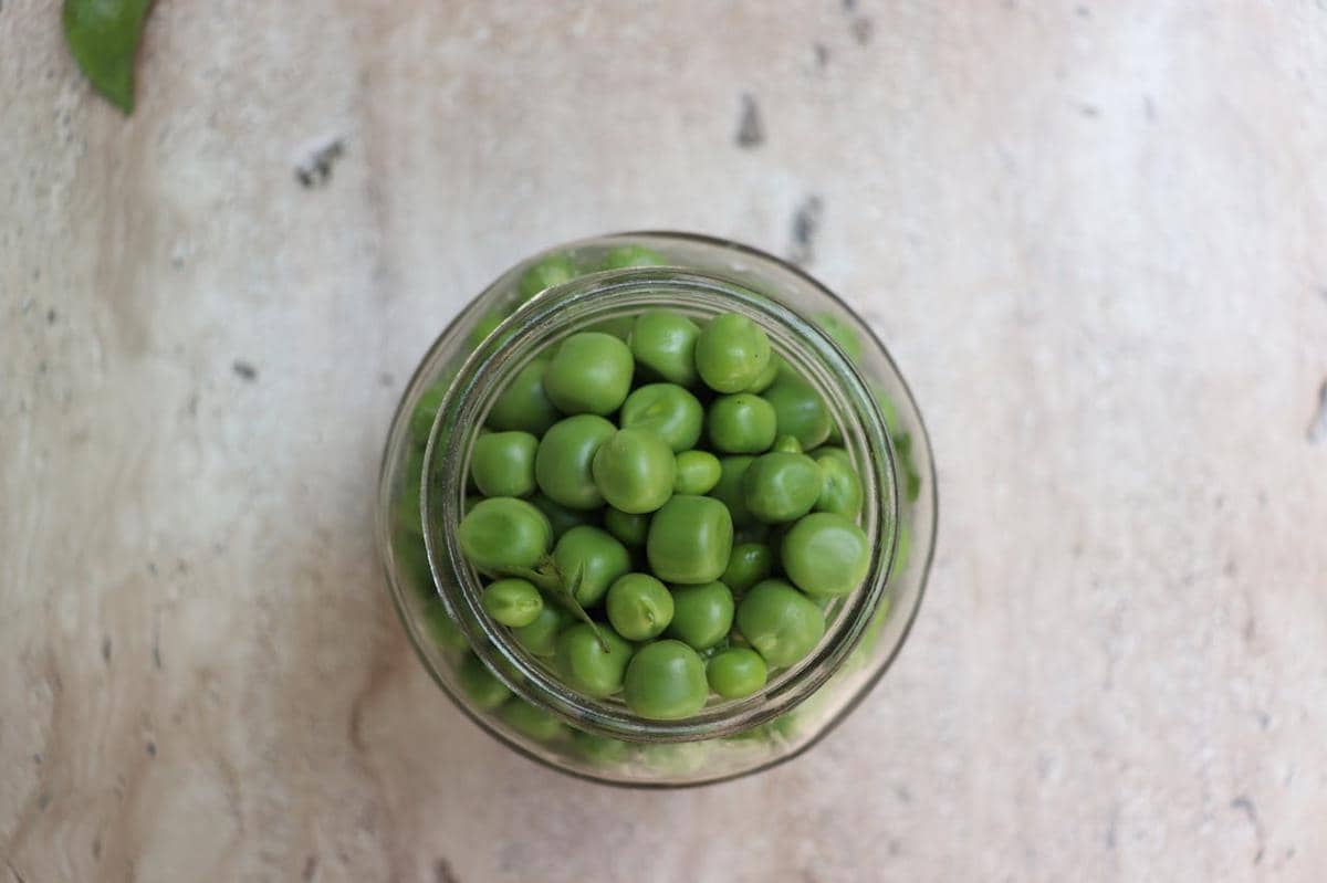 Kacang polong hijau segar di dalam toples kaca dilihat dari atas di atas permukaan meja berwarna terang.