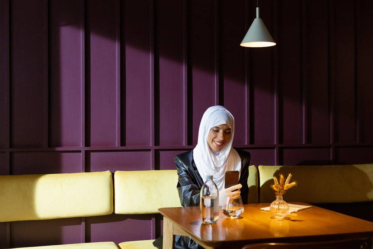 Seorang wanita berhijab tersenyum sambil melakukan video call menggunakan ponsel di meja kafe dengan pencahayaan hangat.