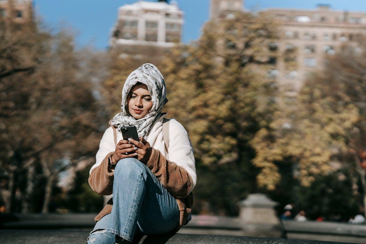Seorang perempuan berhijab duduk di luar ruangan sambil melihat ponsel, dengan latar belakang gedung dan pepohonan.