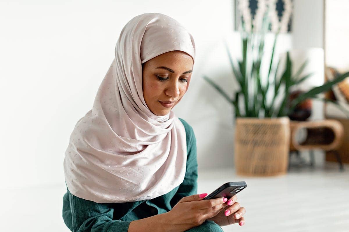 Seorang wanita berhijab duduk sambil memegang ponsel di ruangan terang dengan latar tanaman hias dan perabot rotan.