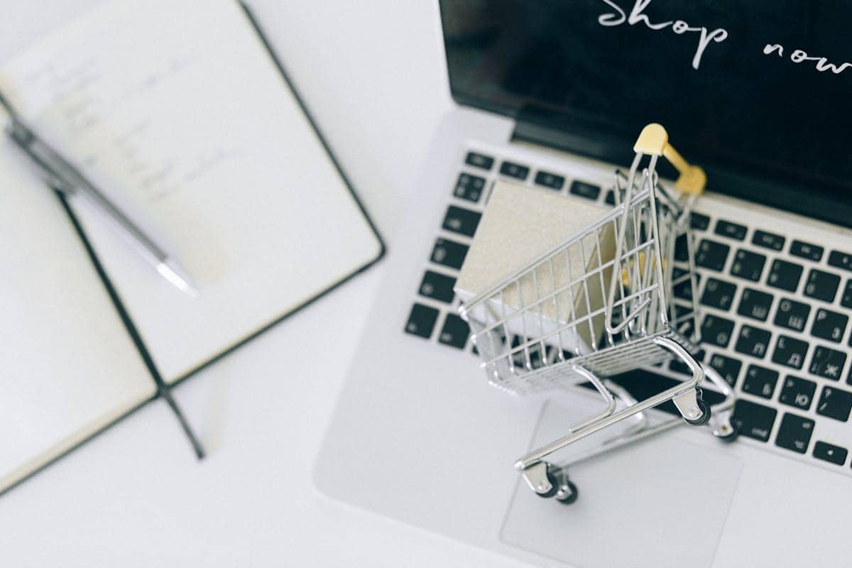 Keranjang belanja mini di atas laptop dengan layar bertuliskan 'shop now' dan buku catatan di sampingnya, menggambarkan konsep belanja online aman.
