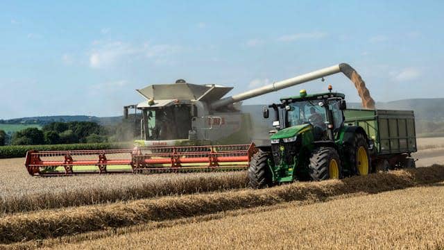 Traktor hijau dan mesin pemanen bekerja di ladang gandum luas saat proses panen berlangsung di bawah langit cerah.