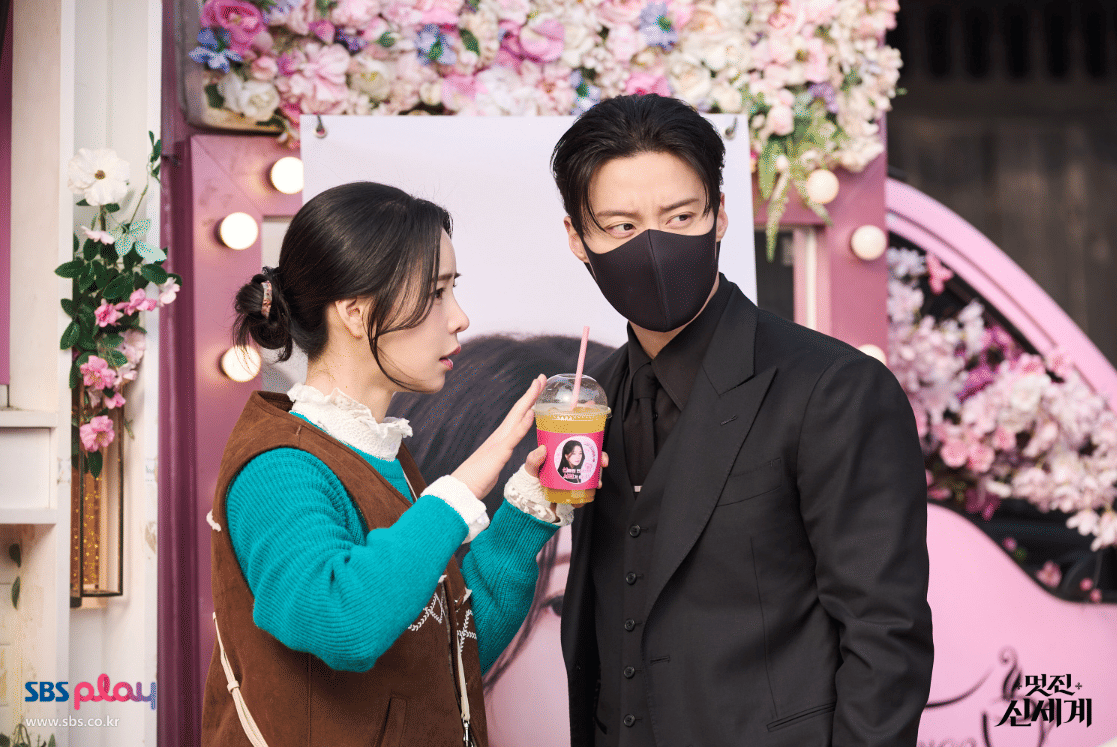 Dua karakter dalam drama My Royal Nemesis berdiri di depan latar bunga berwarna pastel, salah satunya mengenakan masker hitam dan setelan gelap.