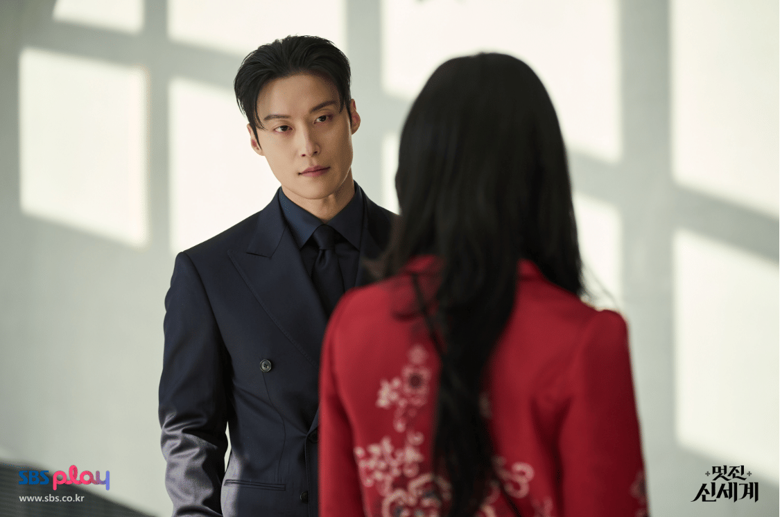 Seorang pria berjas hitam menatap seorang wanita berambut panjang mengenakan pakaian merah dalam adegan drama My Royal Nemesis.