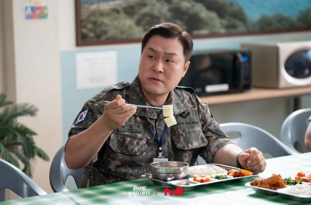 Seorang tentara berseragam militer duduk di ruang makan sambil memegang sumpit dan sepotong makanan di hadapannya terdapat nampan berisi hidangan.