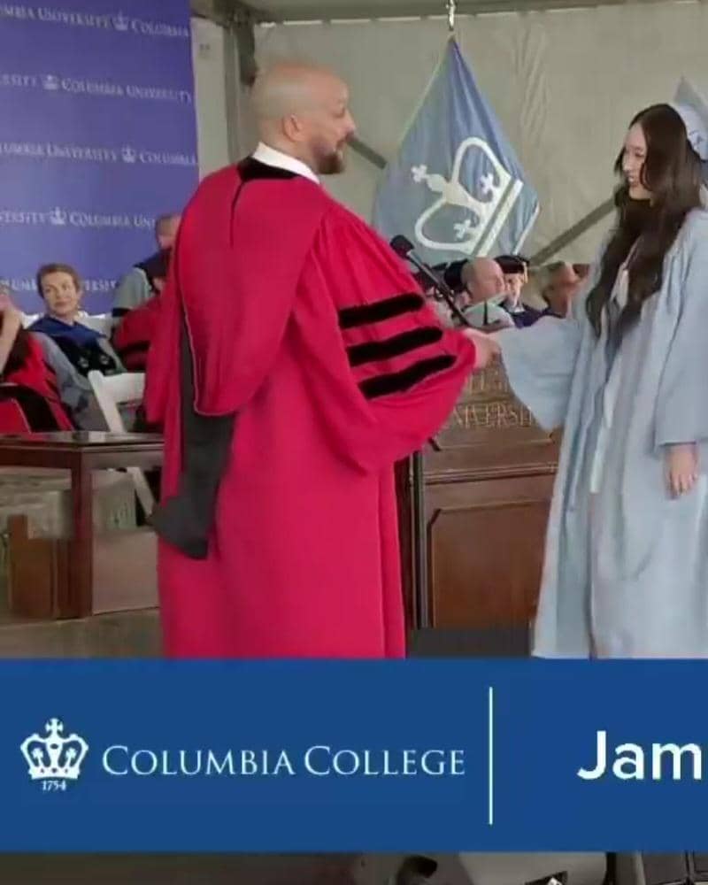 Seorang wisudawan mengenakan toga biru muda berjabat tangan dengan dosen berseragam merah di upacara kelulusan Columbia University.