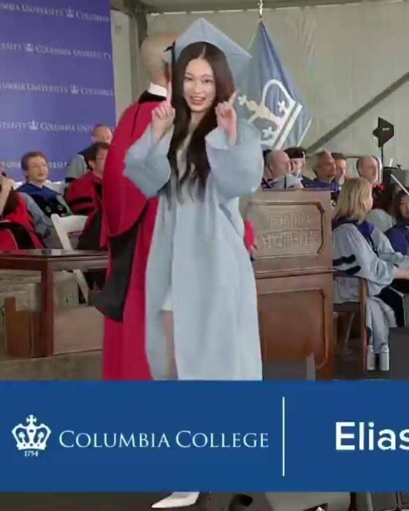 Seorang wisudawati mengenakan toga biru muda dan topi wisuda berdiri di panggung upacara kelulusan Columbia University.