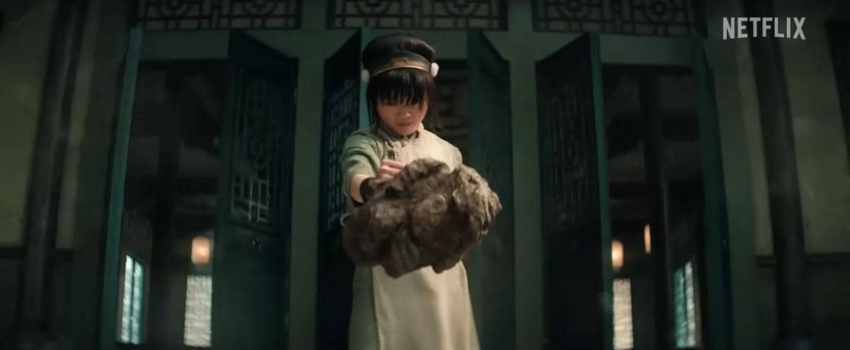 Seorang gadis muda mengenakan pakaian tradisional hijau memanipulasi batu besar di dalam ruangan dengan pintu terbuka di belakangnya.