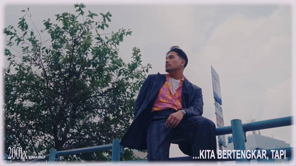 Seorang pria mengenakan jas abu-abu dan kemeja oranye duduk di pagar biru dengan latar pepohonan dan langit cerah.