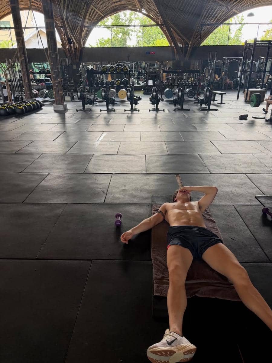 Seorang pria berbaring di lantai gym setelah berolahraga, dengan latar belakang peralatan kebugaran dan atap bambu alami di Lombok.