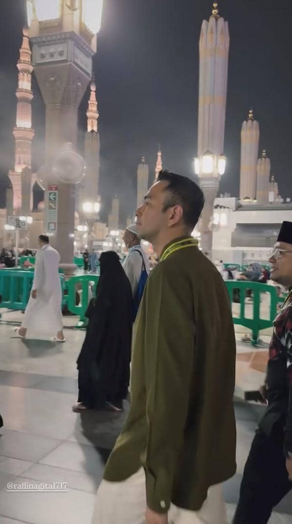 Seorang pria mengenakan pakaian ihram dan jaket hijau berjalan di area Masjid Nabawi pada malam hari bersama jamaah lain.