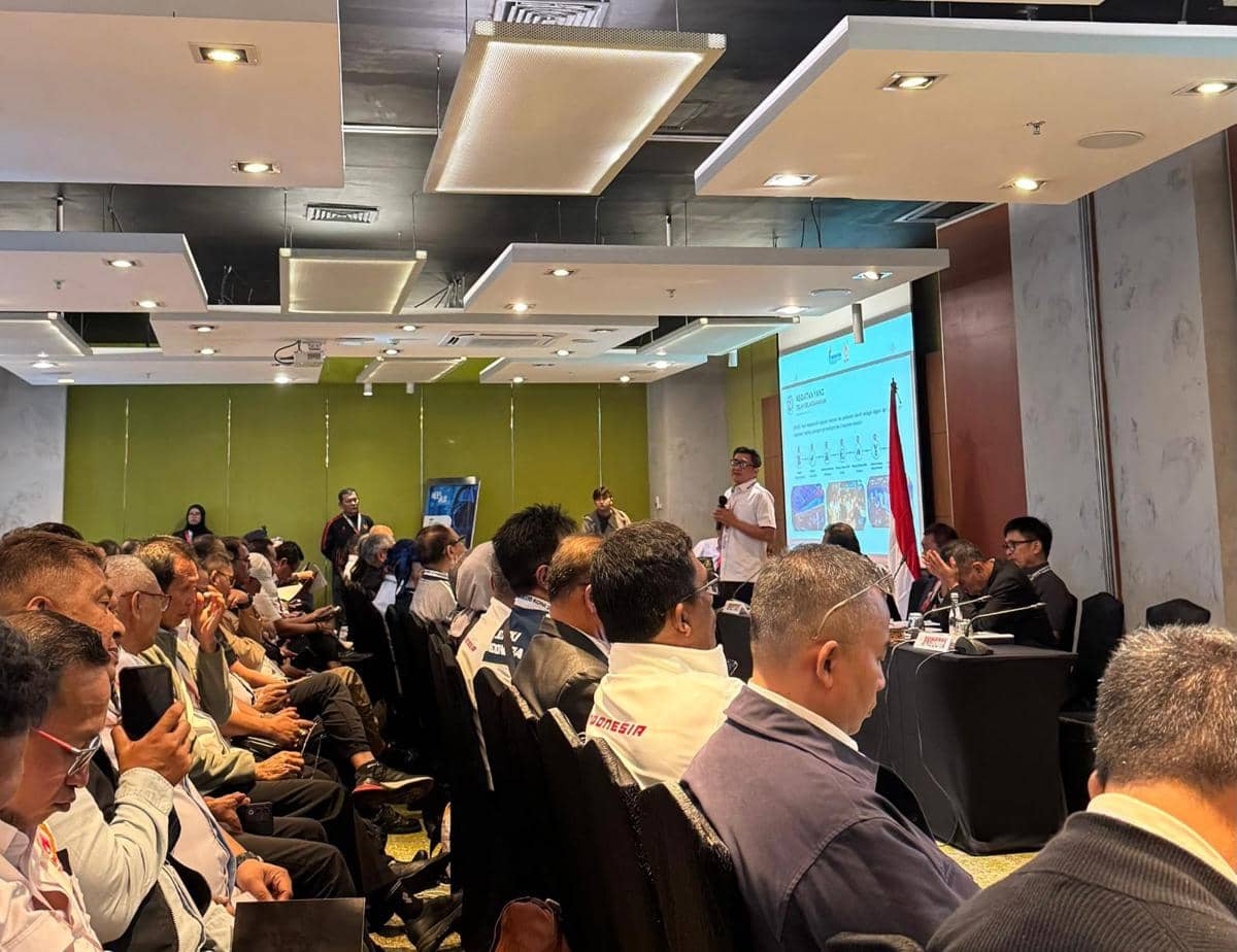 Suasana rapat resmi dengan peserta duduk berbaris mendengarkan presentasi di ruangan konferensi dengan layar proyektor dan bendera Indonesia.