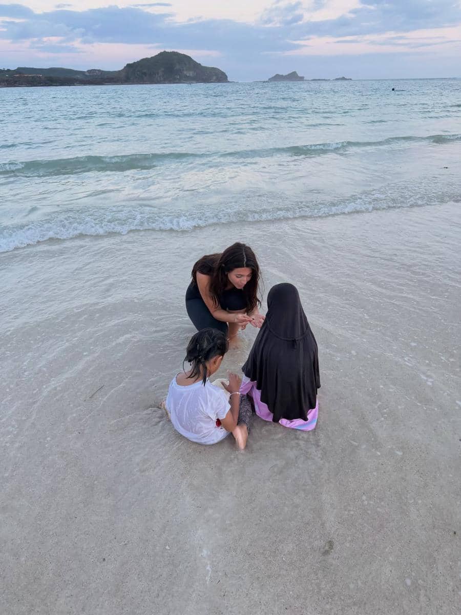 Seorang wanita bersama dua anak kecil duduk di tepi pantai berpasir di Lombok dengan latar laut dan pulau di kejauhan.
