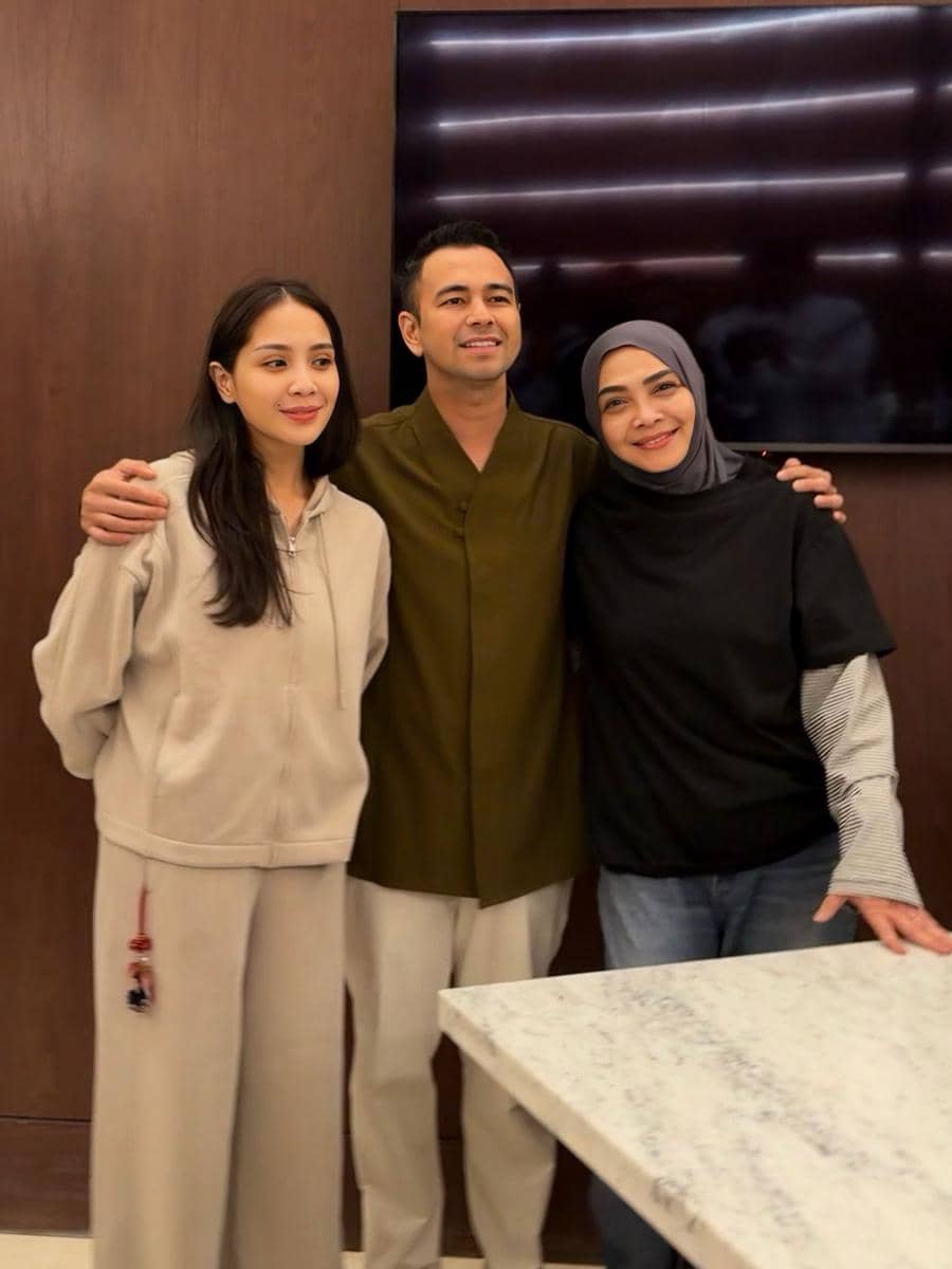 Tiga orang berdiri berdekatan sambil tersenyum di dalam ruangan dengan latar dinding kayu dan meja marmer di depannya.
