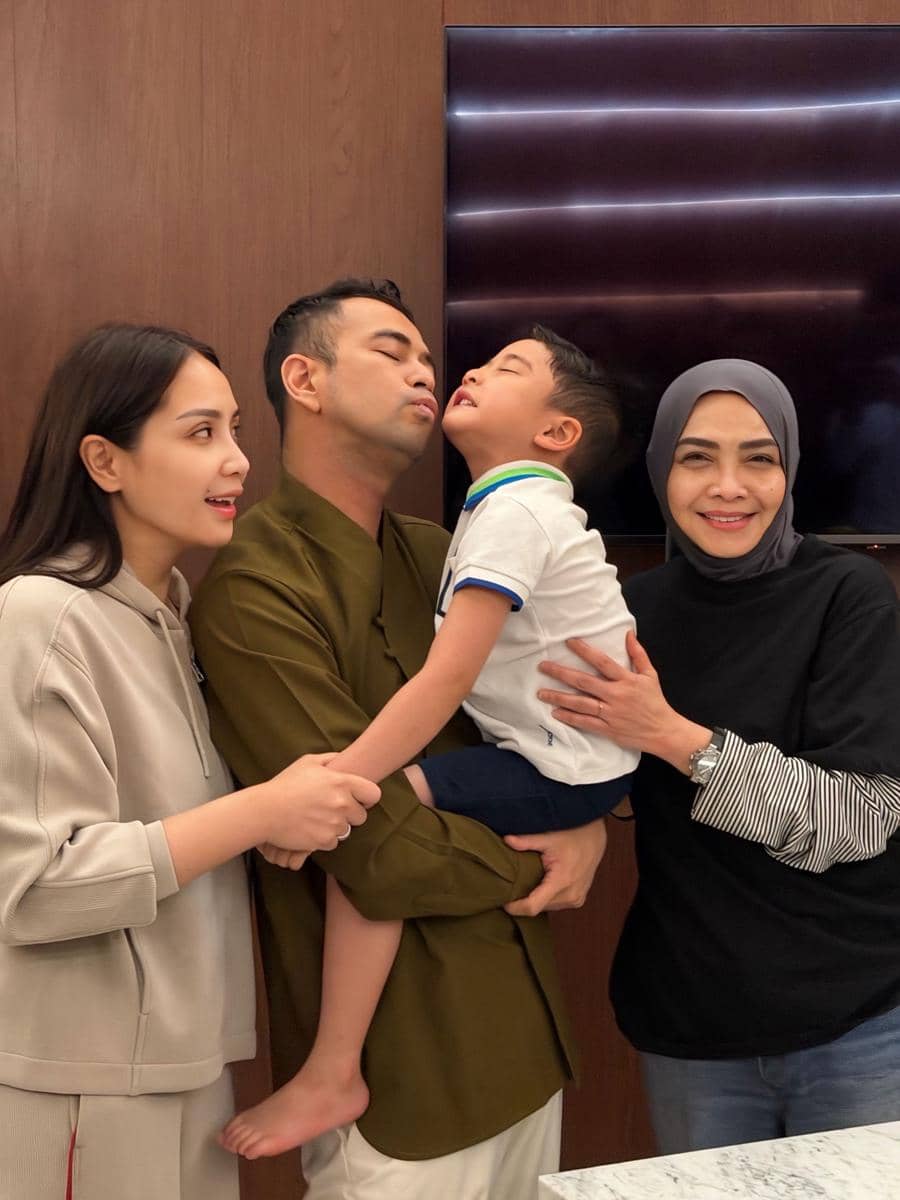 Seorang pria memeluk anak kecil sambil berdiri bersama dua wanita di ruangan dengan dinding kayu dan televisi di belakang mereka.