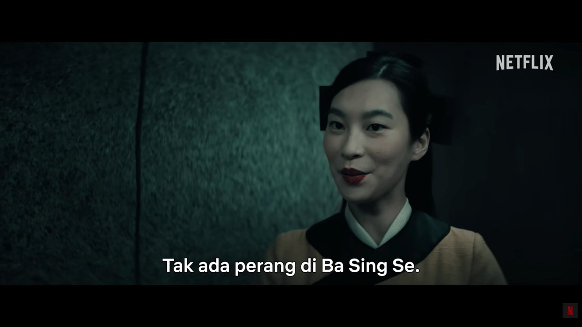 Karakter Joo Dee dalam versi live-action Avatar: The Last Airbender tersenyum dengan latar dinding gelap dan logo Netflix di pojok kanan atas.