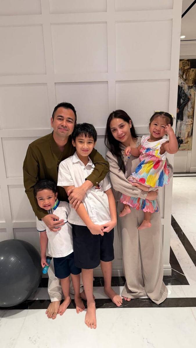 Seorang pria dan wanita berpose bersama tiga anak mereka di dalam ruangan dengan dinding putih, tampak tersenyum hangat ke arah kamera.