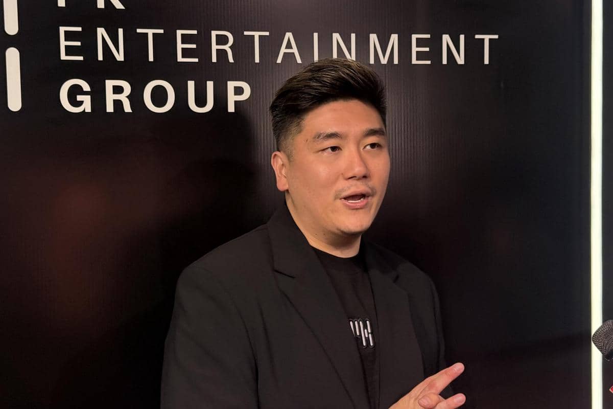 Seorang pria mengenakan jas hitam berbicara di depan latar bertuliskan PK Entertainment Group dengan pencahayaan redup.