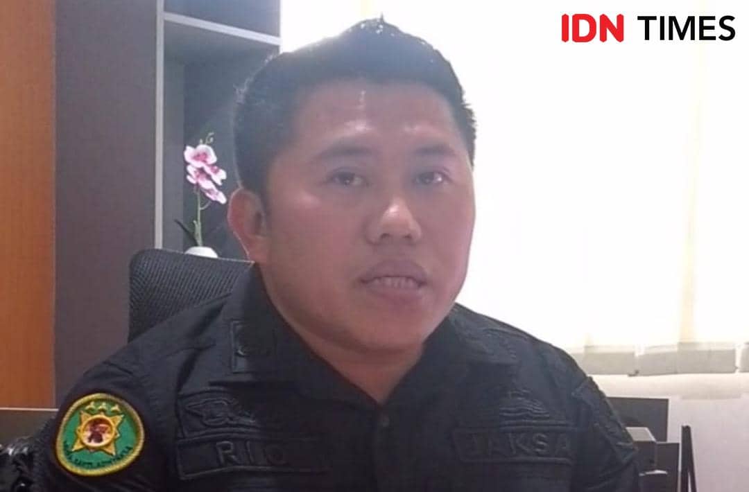 Seorang pria mengenakan seragam hitam dengan lambang Kejaksaan duduk di ruang kantor dengan latar belakang tirai putih.