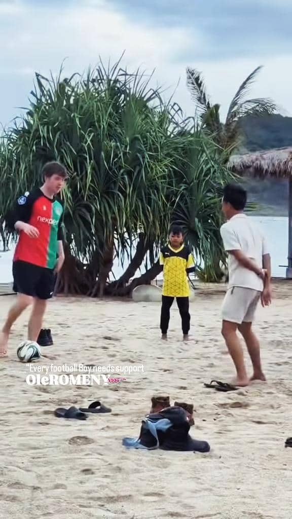 Seorang pria bermain sepak bola di pantai bersama dua anak lokal di Lombok dengan latar pepohonan dan laut di kejauhan.
