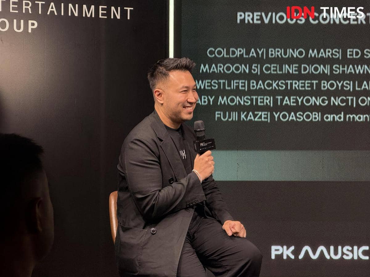 Seorang pria mengenakan setelan hitam sedang duduk sambil memegang mikrofon di acara PK Entertainment Group.