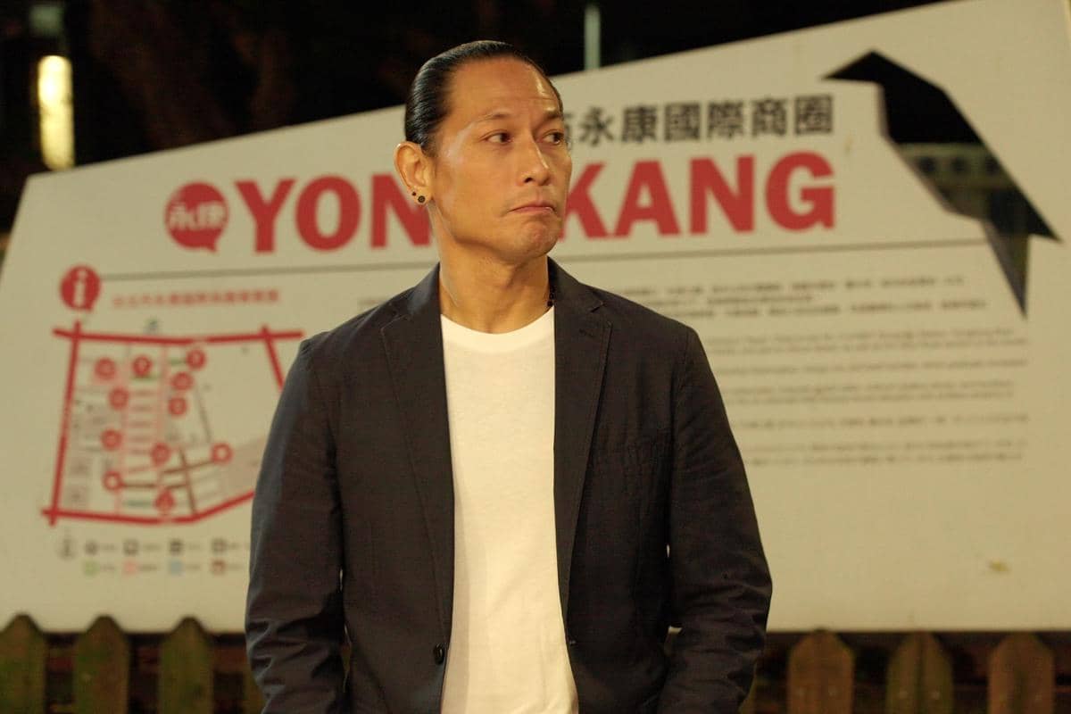 Seorang pria berjaket hitam dan kaus putih berdiri di depan papan bertulisan YONG KANG dengan latar malam hari.