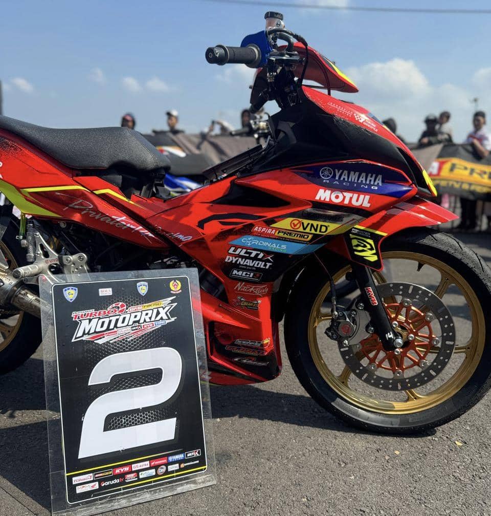 Motor balap berwarna merah dengan stiker sponsor Motul dan Yamaha Racing diparkir di lintasan bersama papan nomor dua.