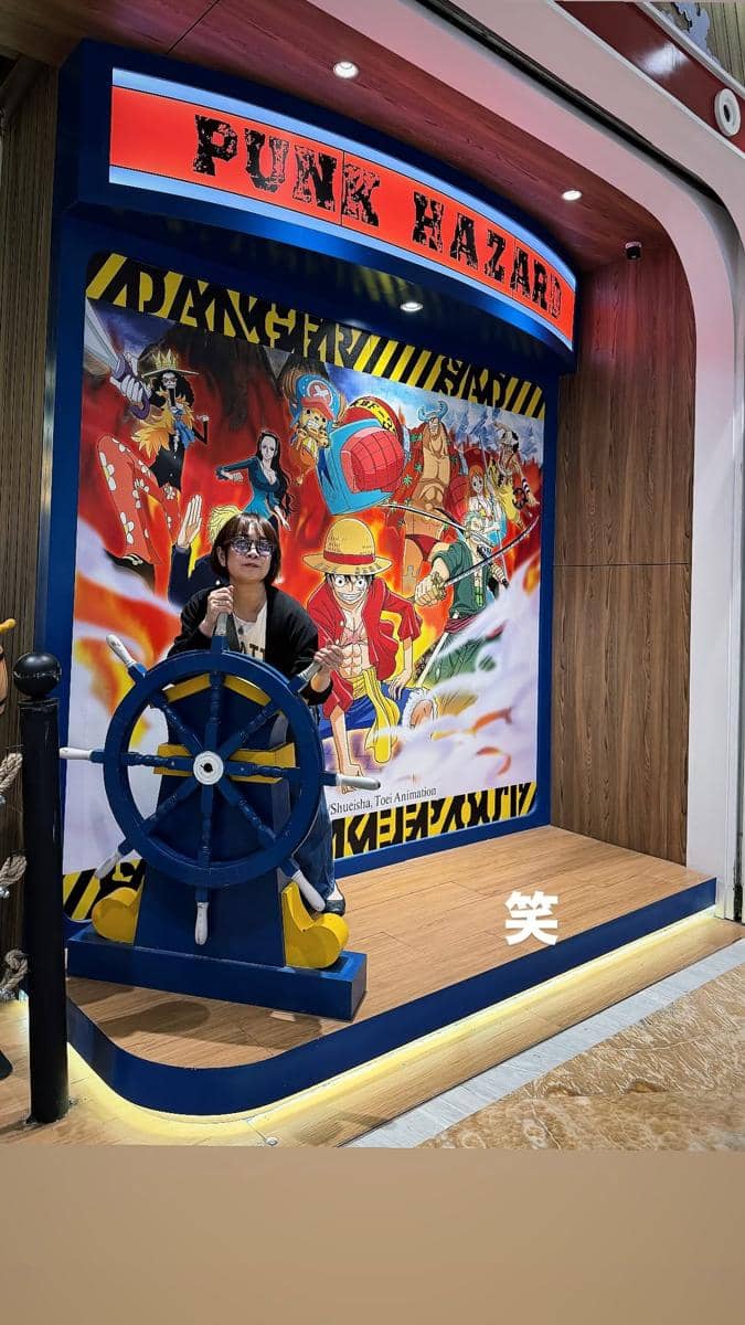 Seorang perempuan berpose di depan mural bertema One Piece dengan tulisan Punk Hazard sambil memegang kemudi kapal di area dalam ruangan.