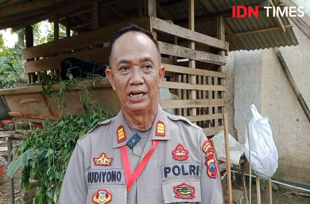 Seorang anggota polisi berseragam berdiri di depan kandang ternak dengan latar tumpukan hijauan pakan dan bangunan kayu sederhana.