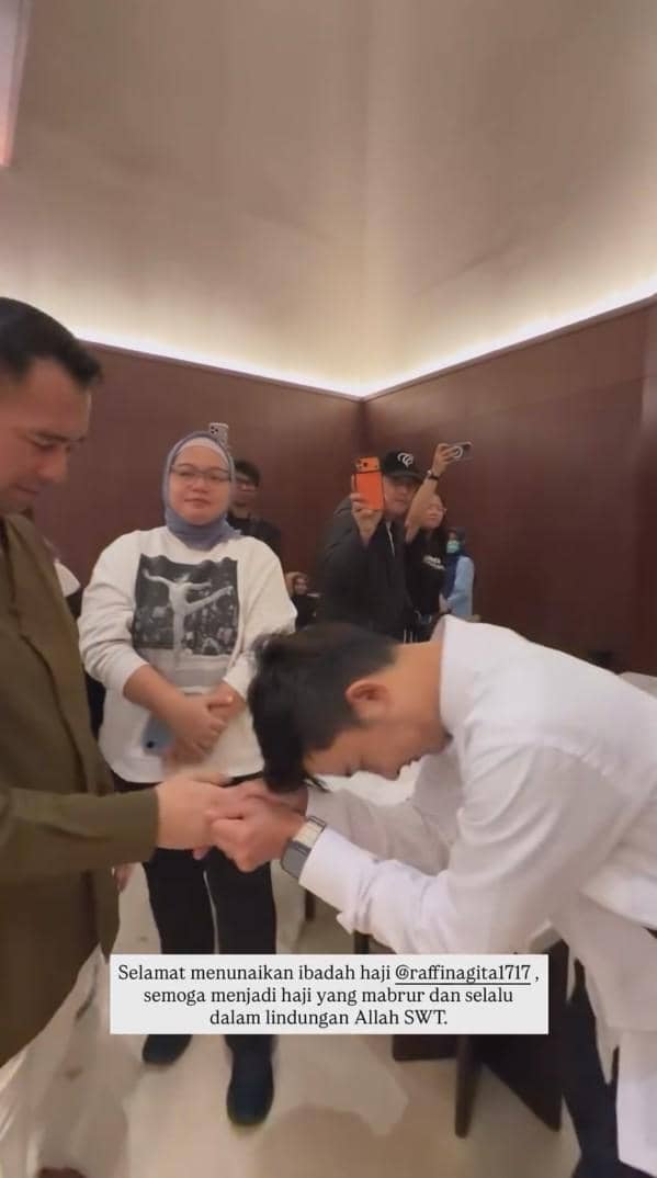 Seorang pria berpakaian putih mencium tangan pria lain sebelum berangkat haji, disaksikan beberapa orang yang merekam momen tersebut.