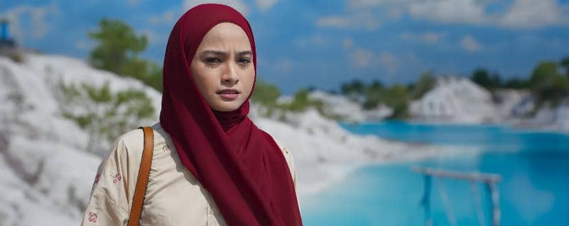 Seorang perempuan berhijab merah berdiri di tepi danau biru dengan latar perbukitan kapur di bawah langit cerah.