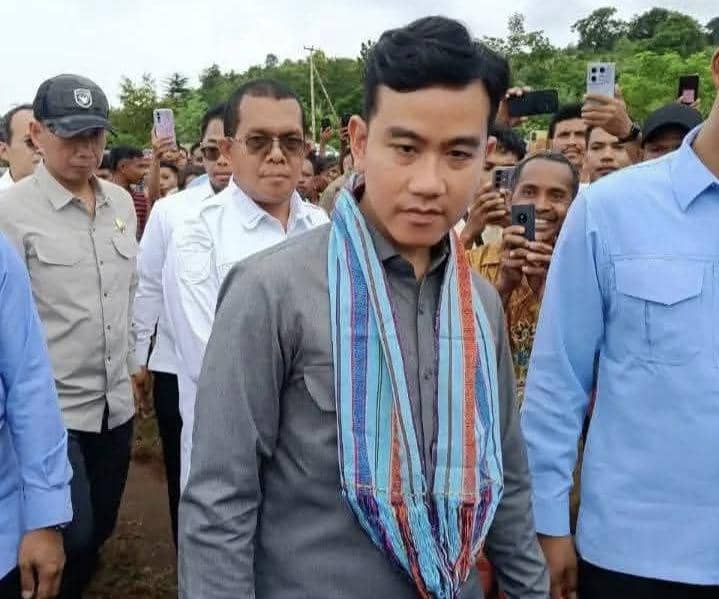 Seorang pria mengenakan kemeja abu-abu dan selendang tenun tradisional berdiri di antara rombongan dan warga di Amfoang, NTT.