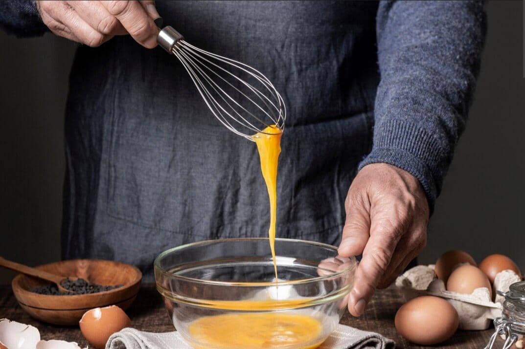 Seseorang mengenakan sweater biru sedang mengocok telur dengan whisk di atas mangkuk kaca di meja dapur dengan beberapa telur di sekitarnya.