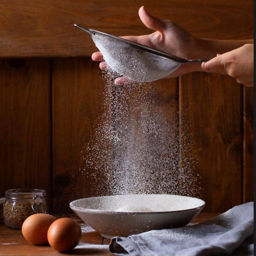 Tangan sedang mengayak tepung ke dalam mangkuk di atas meja kayu dengan dua butir telur dan toples kecil di sekitarnya.