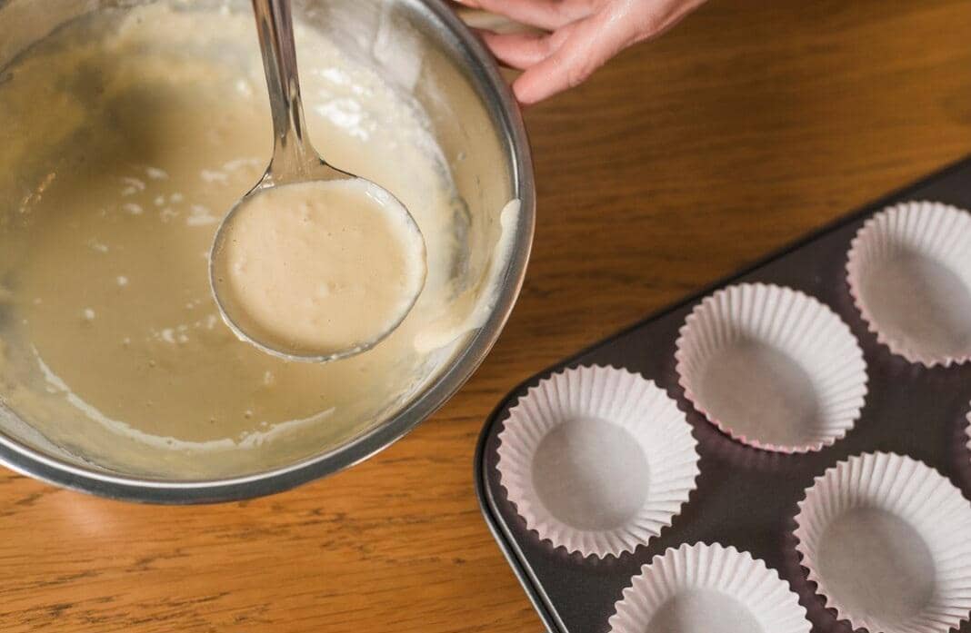 Adonan kue cair di dalam mangkuk logam sedang disendok untuk dituangkan ke cetakan muffin kertas di atas meja kayu.