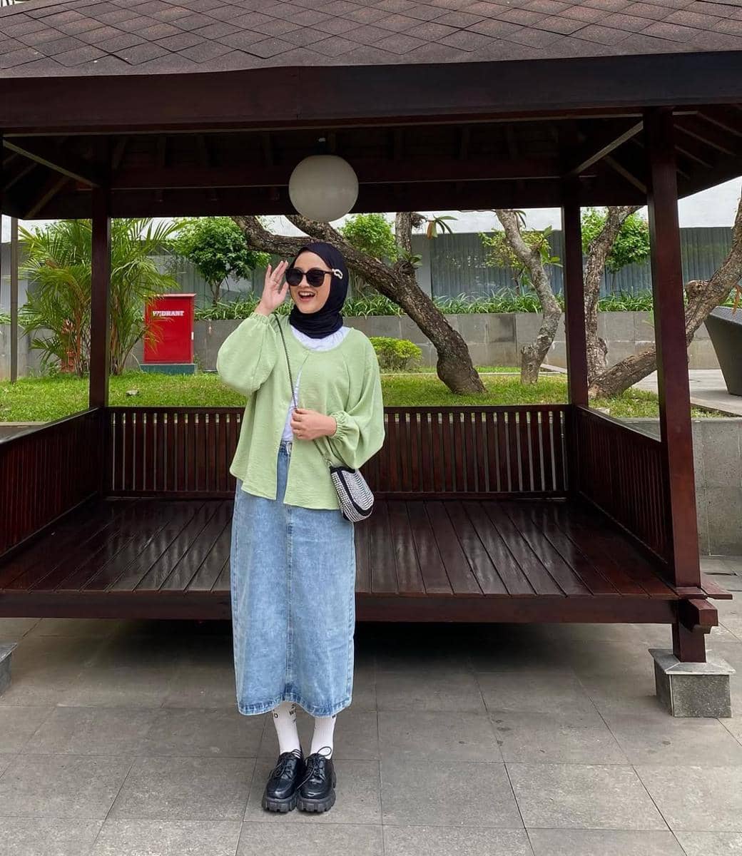 Seorang wanita berhijab mengenakan cardigan hijau muda, rok denim panjang, dan sepatu hitam berpose di depan gazebo kayu di area taman.