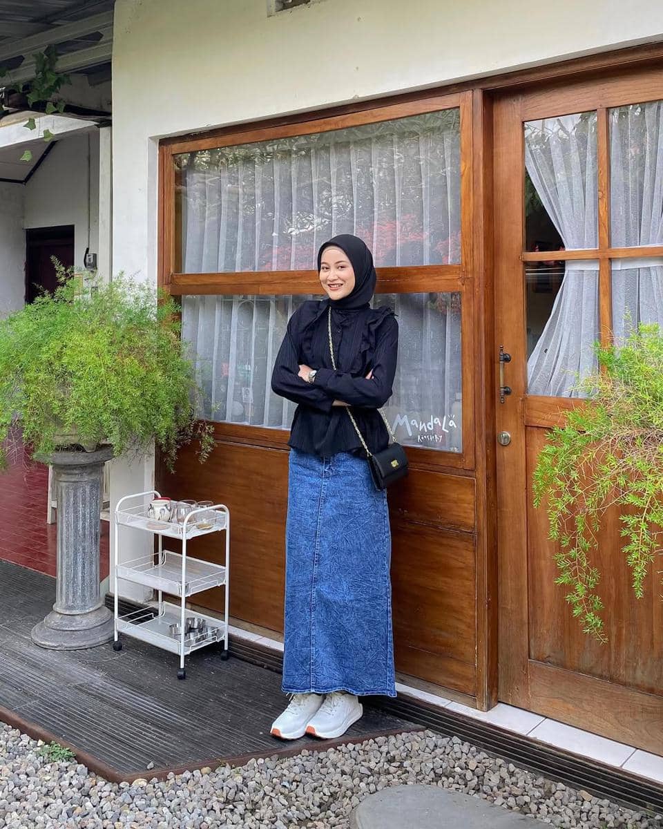 Seorang wanita berhijab mengenakan atasan hitam dan rok denim panjang berpose di depan pintu kayu dengan latar tanaman hijau.