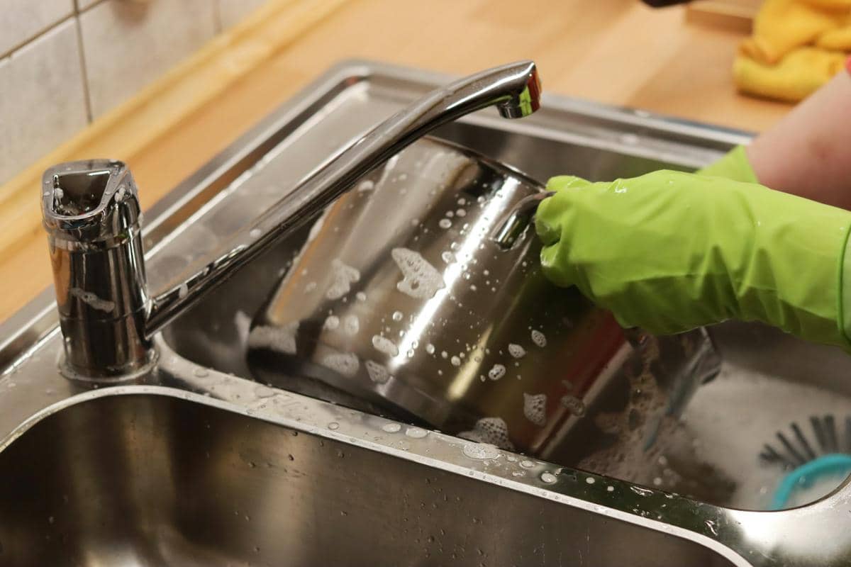 Seseorang mengenakan sarung tangan karet hijau sedang mencuci panci stainless di wastafel dapur dengan air dan sabun.