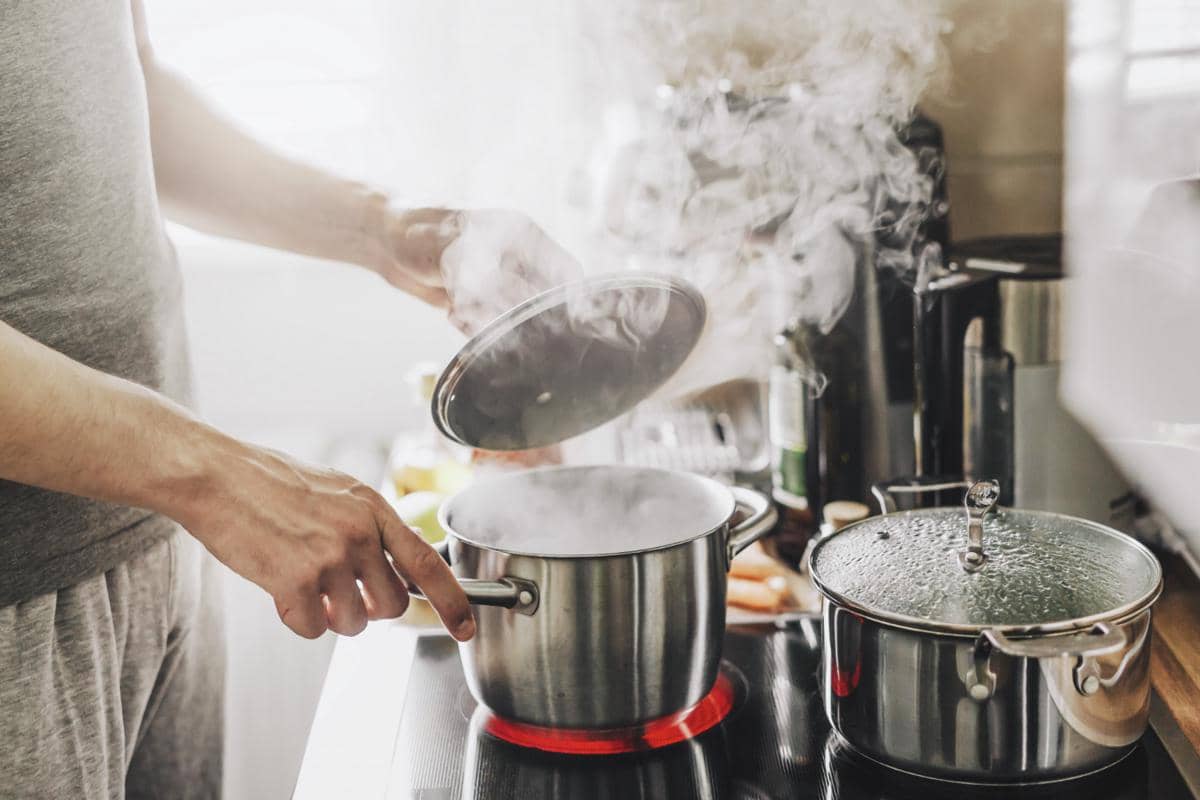 Seseorang membuka tutup panci berisi makanan yang sedang dikukus di atas kompor dengan uap panas mengepul di dapur.