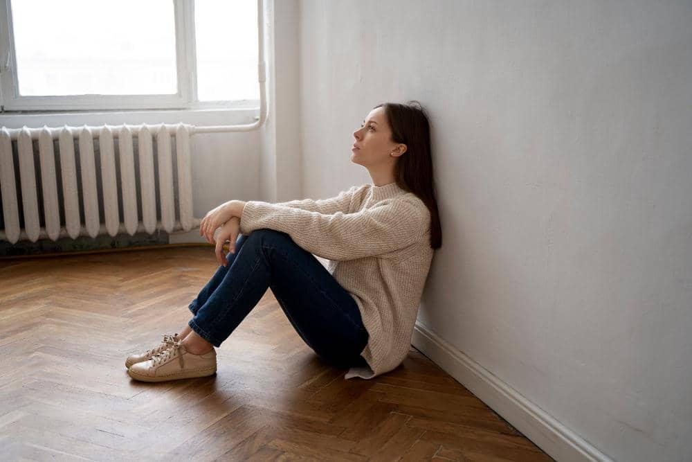 Seorang perempuan duduk bersandar di dinding kamar dengan ekspresi termenung, mengenakan sweter krem dan celana jeans biru.