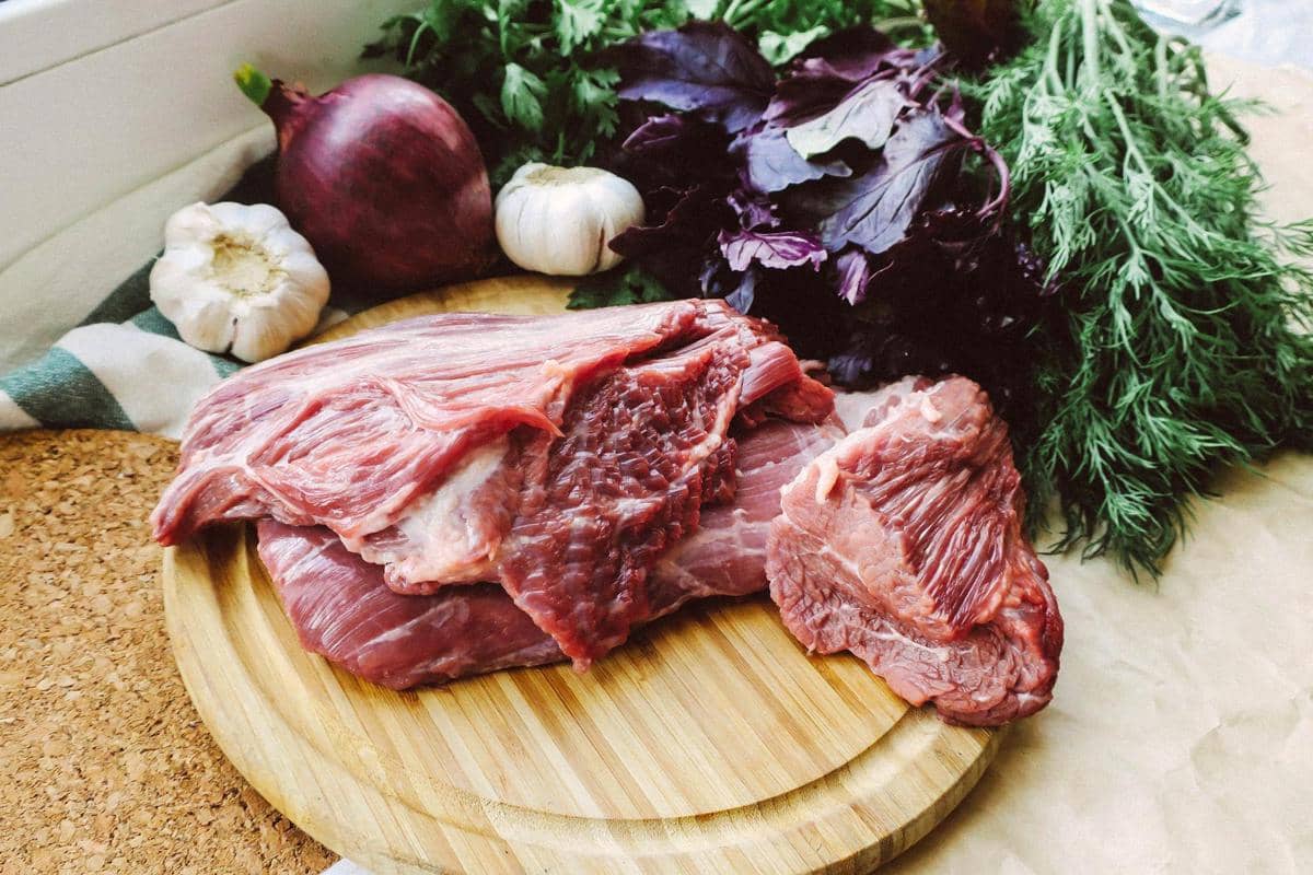 Potongan daging kambing segar di atas talenan kayu dengan bawang putih, bawang merah, dan sayuran hijau di sekitarnya.