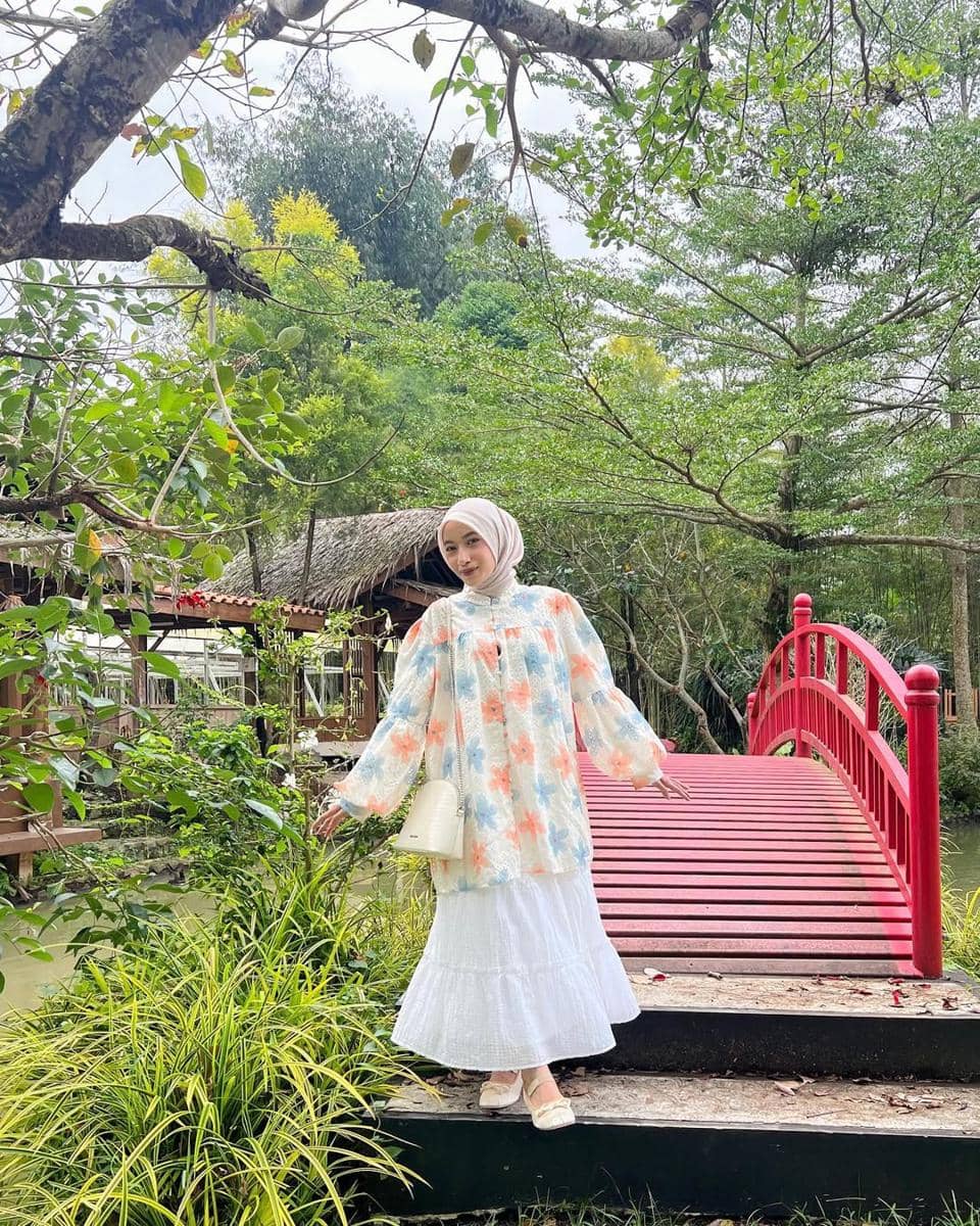 Seorang wanita berhijab berpose di atas jembatan merah di taman hijau, mengenakan tunik bermotif bunga dan rok putih panjang.